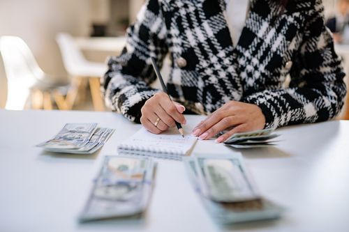 Free A woman in a plaid blazer writes in a notepad with cash spread on a white table, focusing on financial planning. Stock Photo