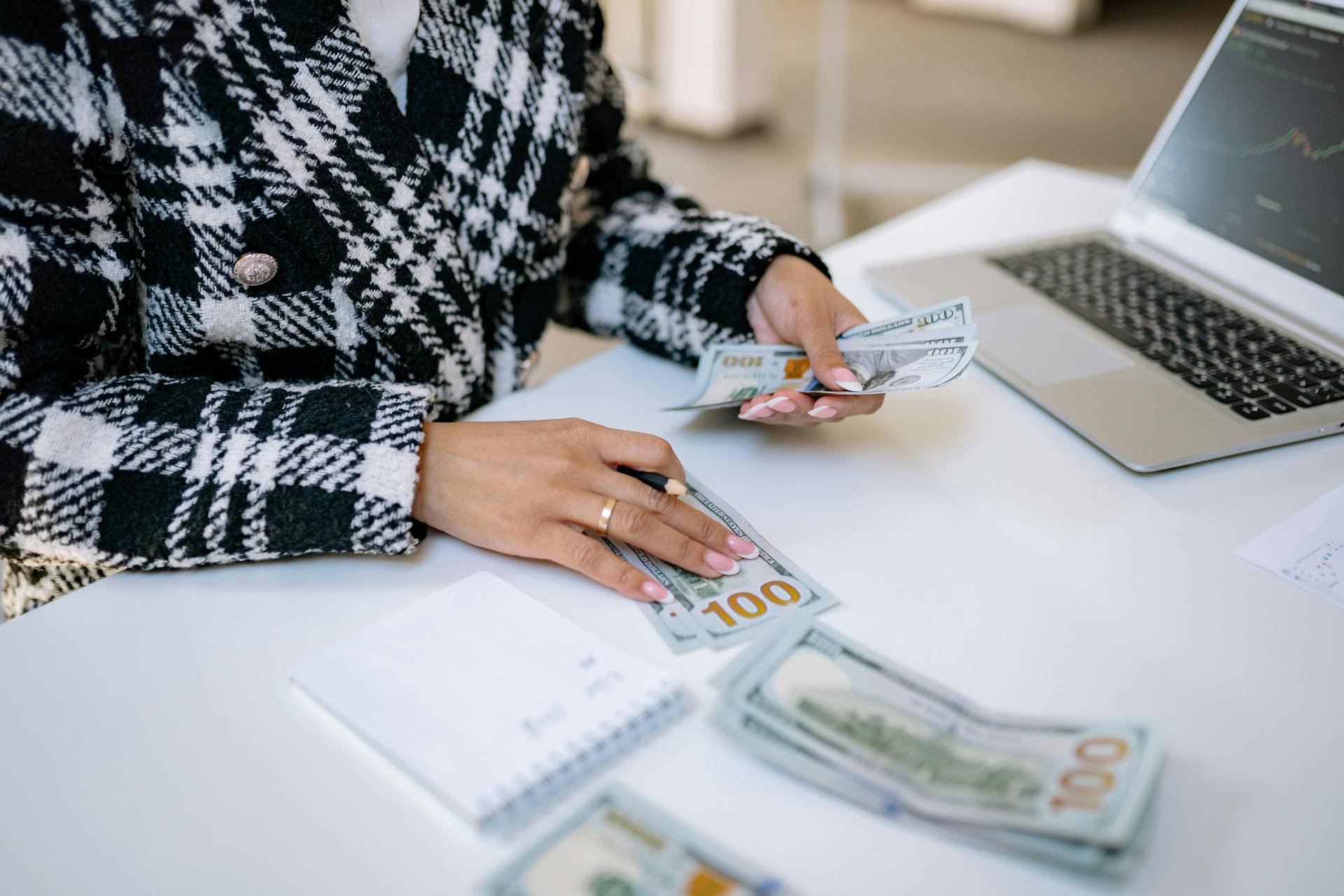 Close-up of a woman counting hundred-dollar bills at a desk with a laptop, focusing on finance and work.