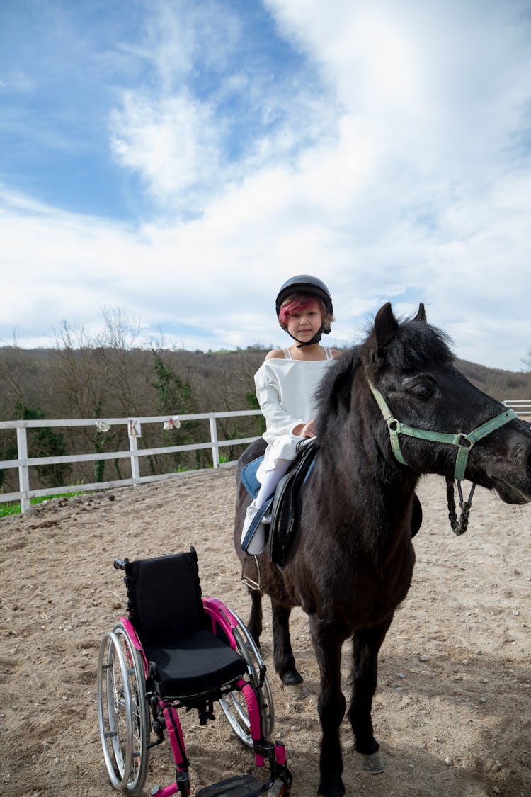 Smiling Girl On Black Pony With Wheelchair Near