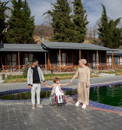 Full body of happy father and mother in hijab touching hands of little daughter in wheelchair near pool of garden