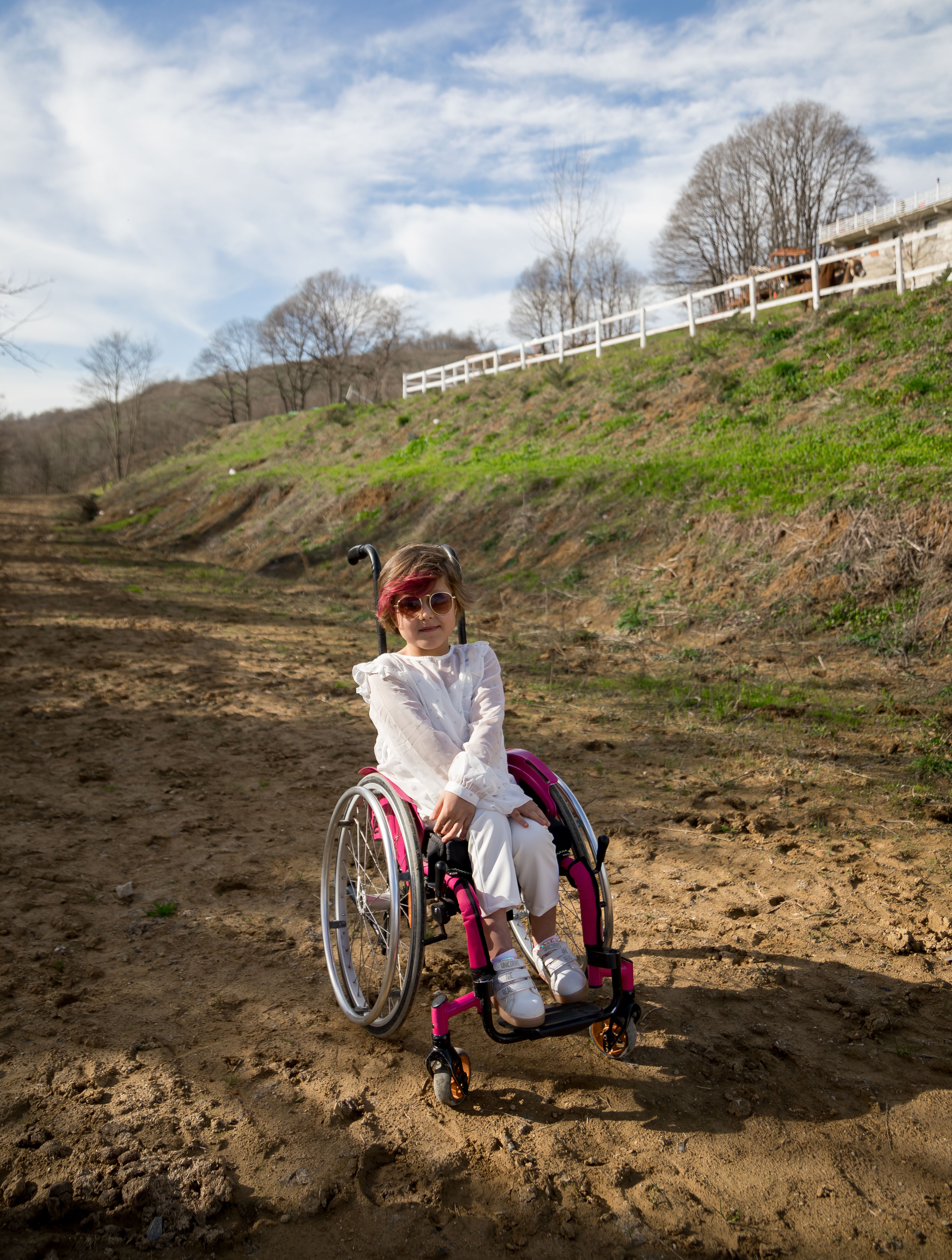 Cute girl in pink wheelchair in park with fresh green grass under peaceful blue cloudy sky