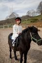 Adorable happy girl smiling and looking at camera on black pony in paddock under cloudy blue sky
