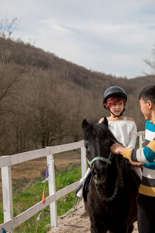 Free Cute girl in helmet talking with crop anonymous male while riding pony in paddock in cloudy weather Stock Photo