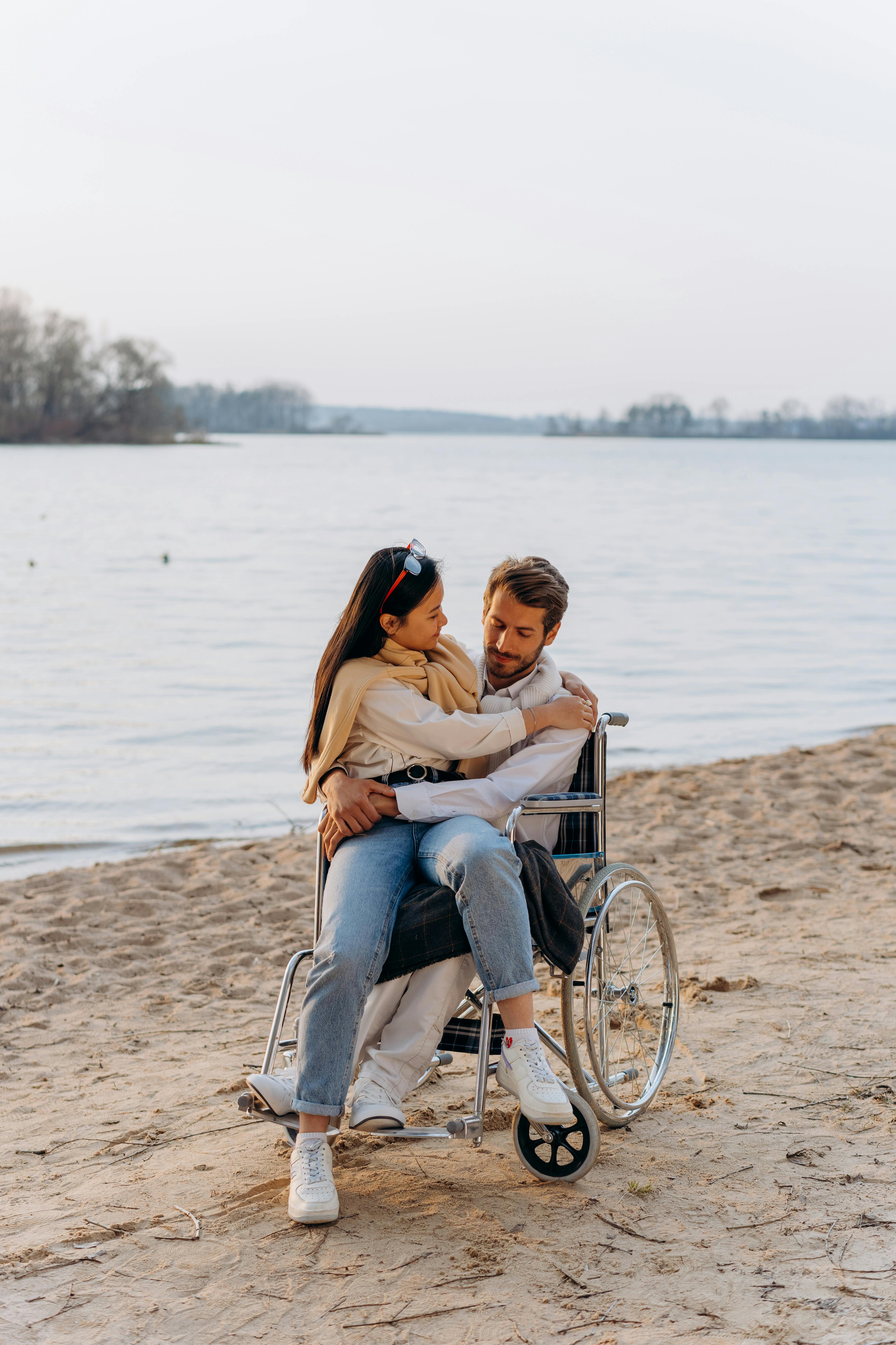 A couple enjoying a tender moment by the beach, with one seated in a wheelchair, showcasing love and accessibility.