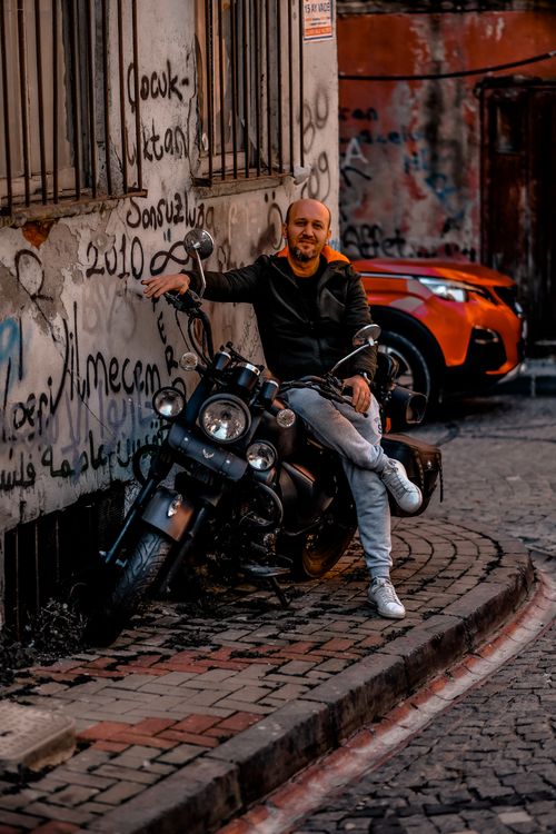 Free A man casually sits on a motorcycle against a graffiti-covered wall in an urban street with a car in the background. Stock Photo