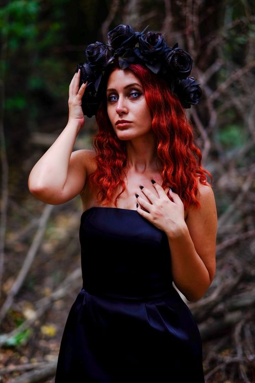 Free Elegant woman with red hair wearing a black dress and flower crown in a forest setting. Captivating and mysterious. Stock Photo