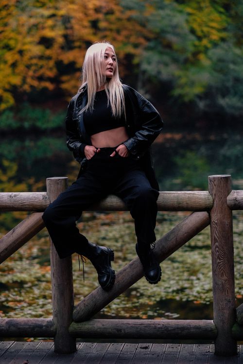 Free Stylish woman in black leather jacket sitting on a wooden fence by a lake in autumn. Stock Photo