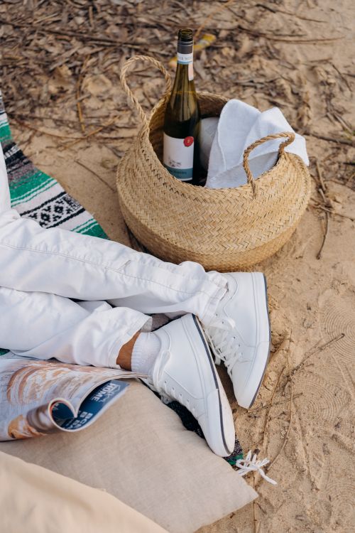 Free Beach picnic scene with woven basket, wine bottle, and white sneakers on sandy ground. Stock Photo