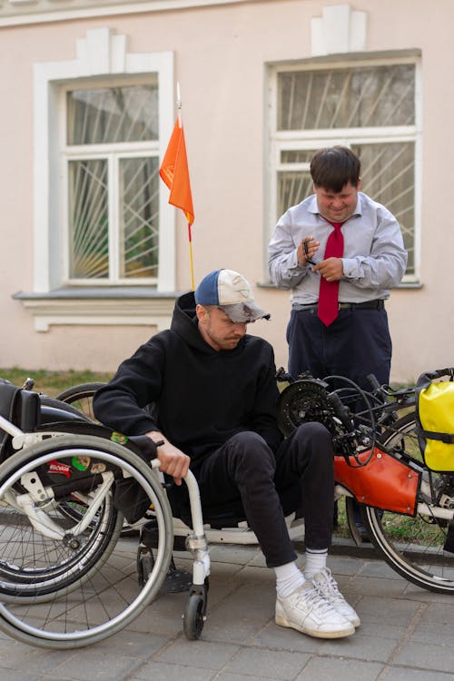 A Man in Blue Long Sleeves Standing Near a Man Sitting on a Handcycle