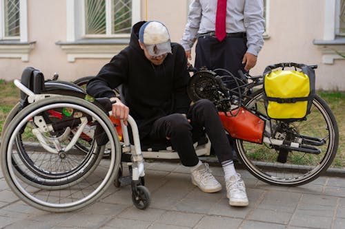 Free A man transferring from a wheelchair to a handcycle on an urban sidewalk, assisted by another person. Stock Photo