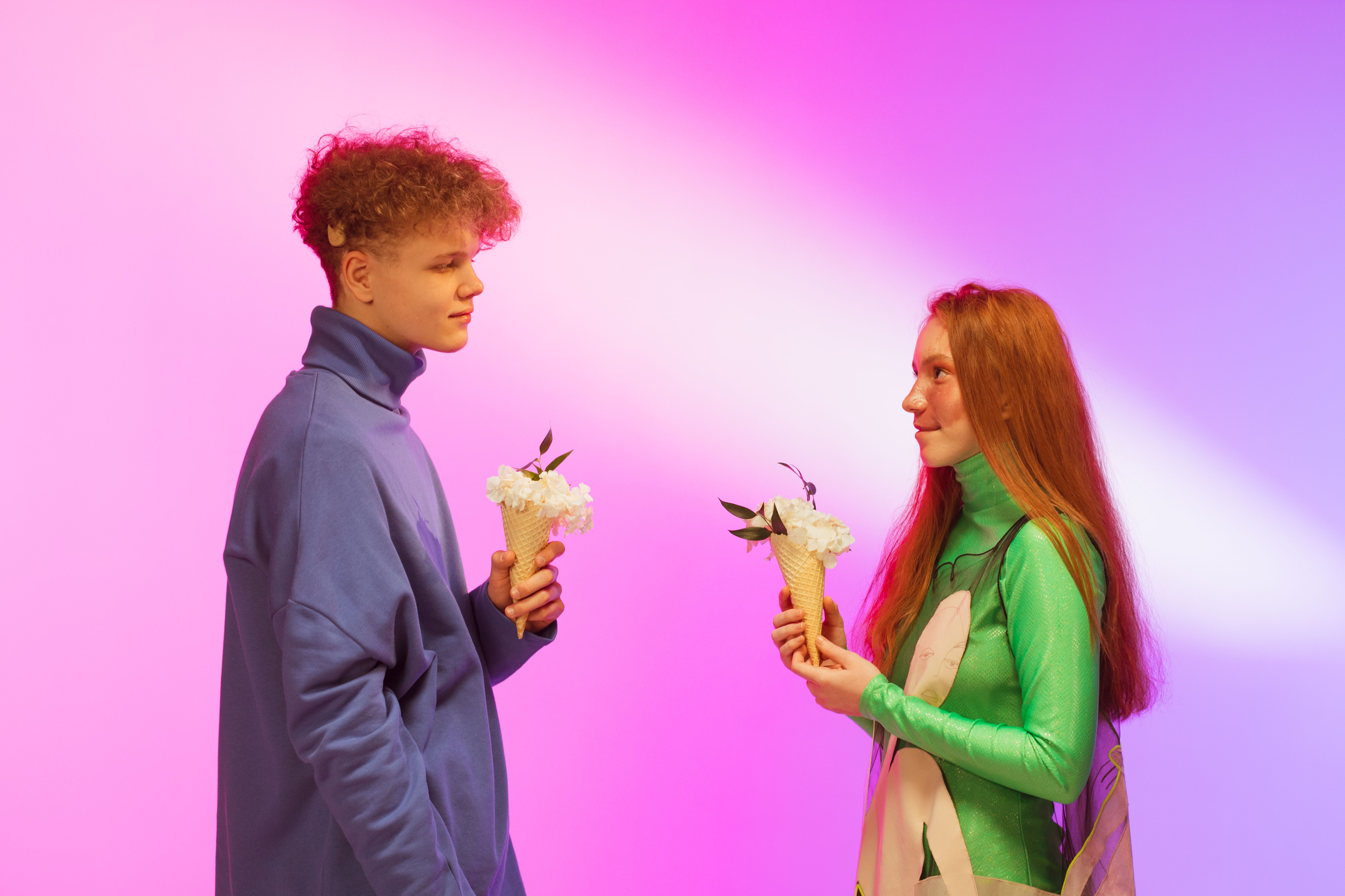 Two young adults gaze at each other, holding floral cones, against a vibrant pink and purple backdrop.