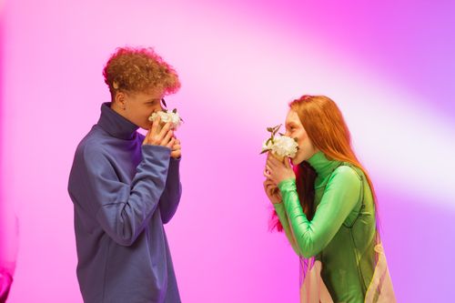 Free A man and woman enjoy smelling flowers against a vibrant pink studio backdrop. Stock Photo