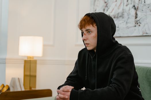 Teenager with bruises sits indoors, deep in thought, wearing a black hoodie.