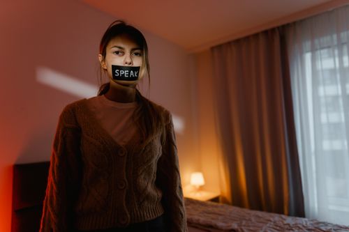 Free A woman stands in a dimly lit bedroom with tape reading 'Speak' over her mouth, symbolizing silence and oppression. Stock Photo