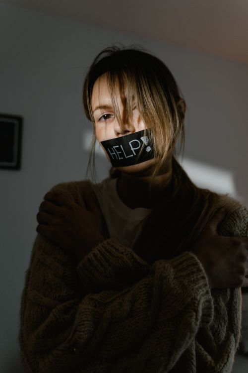 Free A woman with 'help' tape over mouth, expressing distress and urgency indoors. Stock Photo