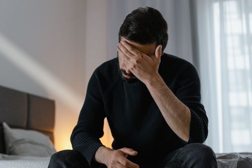 Free A man in distress sits on a bed indoors, covering his face with his hand. Stock Photo