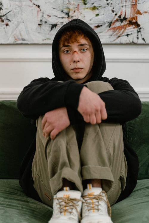 Free A teenage boy wearing a black hoodie sits on a couch, looking introspective. Stock Photo
