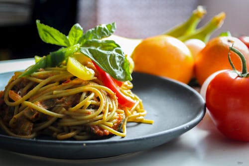 Паста с овощным блюдом на серой тарелке рядом с фруктами помидора на белом столе