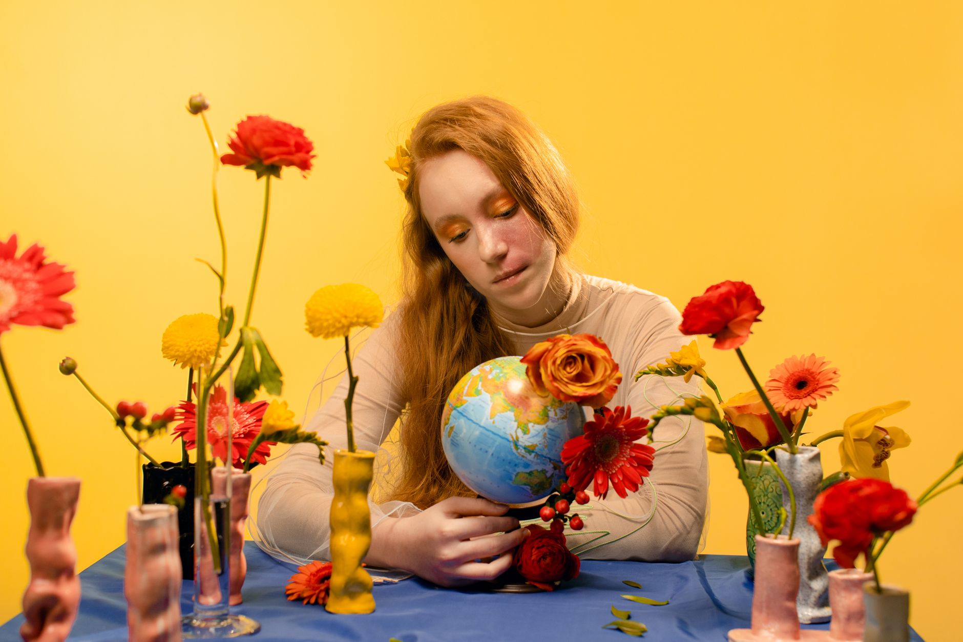 A teenage girl with red hair holding a globe surrounded by colorful flowers in vases, set against a bright yellow background.