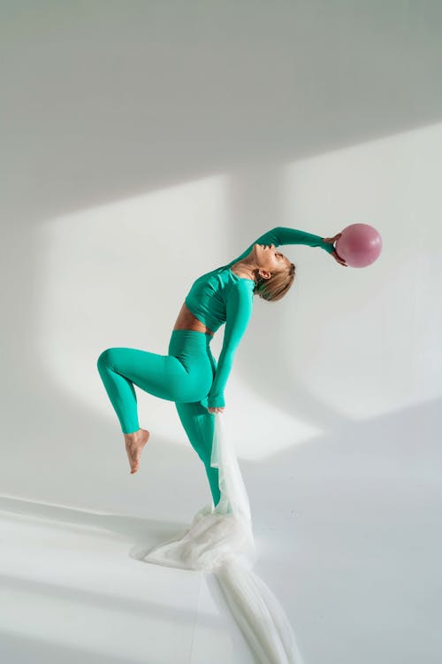 Full body side view of determined fit female gymnast doing exercise with gymnastic ball and bending back while training on white background with leg raised