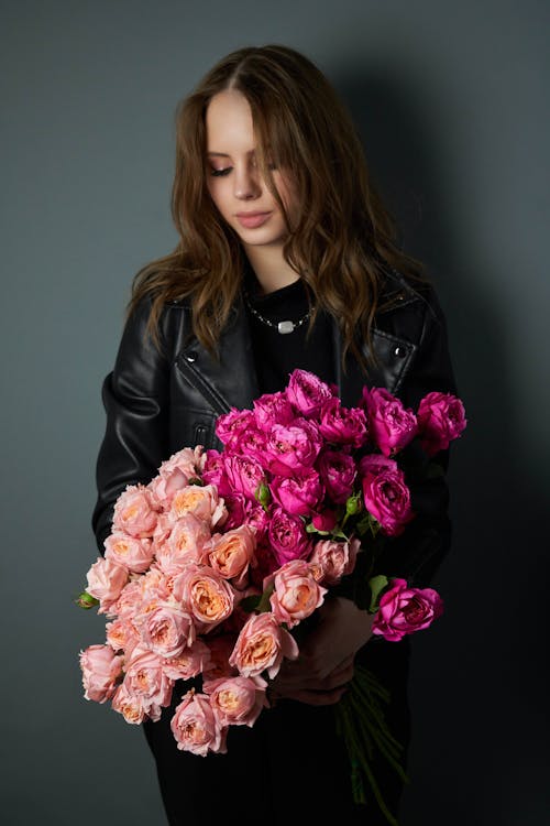 Free Young lady wearing black leather jacket while standing with bouquet of blossoming flowers and closed eyes in light studio on gray background Stock Photo