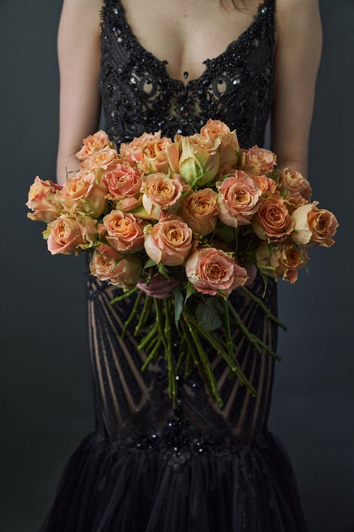 Free Crop anonymous lady wearing black stylish dress while standing with bouquet of blooming flowers on gray background in bright studio Stock Photo