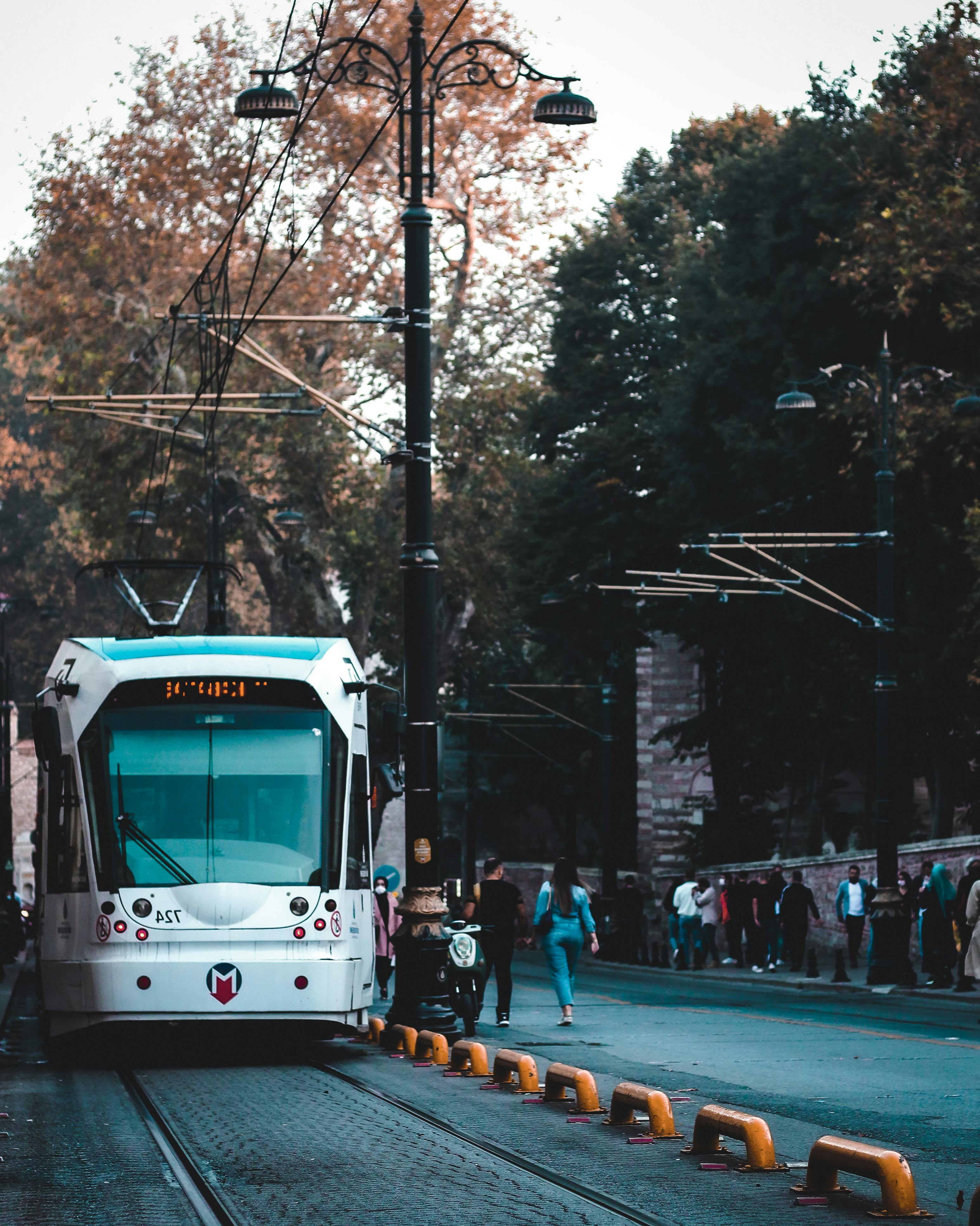 Free A sleek tram traverses a city street lined with trees and pedestrians on a bustling day. Stock Photo