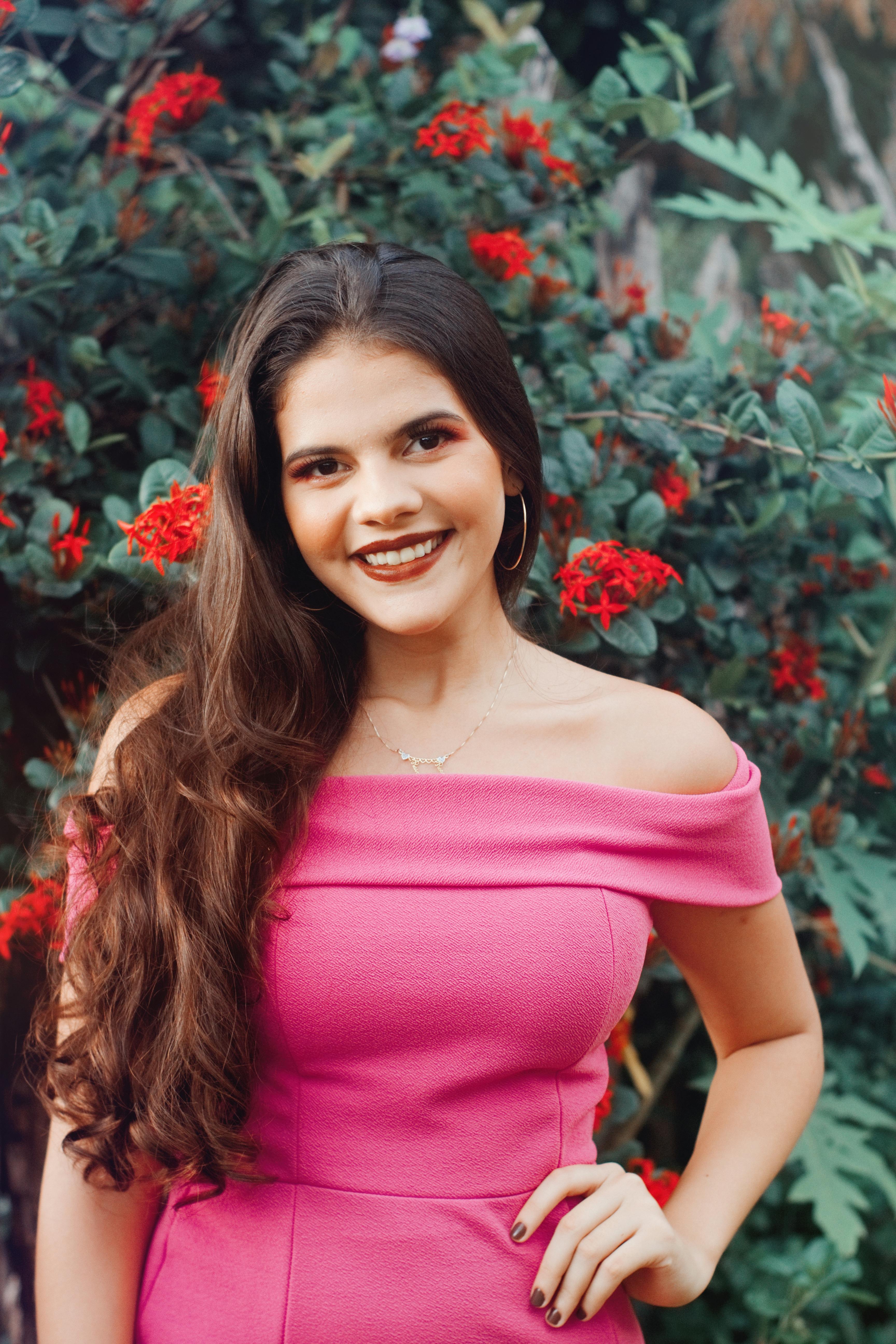Portrait of a smiling woman with long brown hair in a pink dress, set against vibrant red flowers.