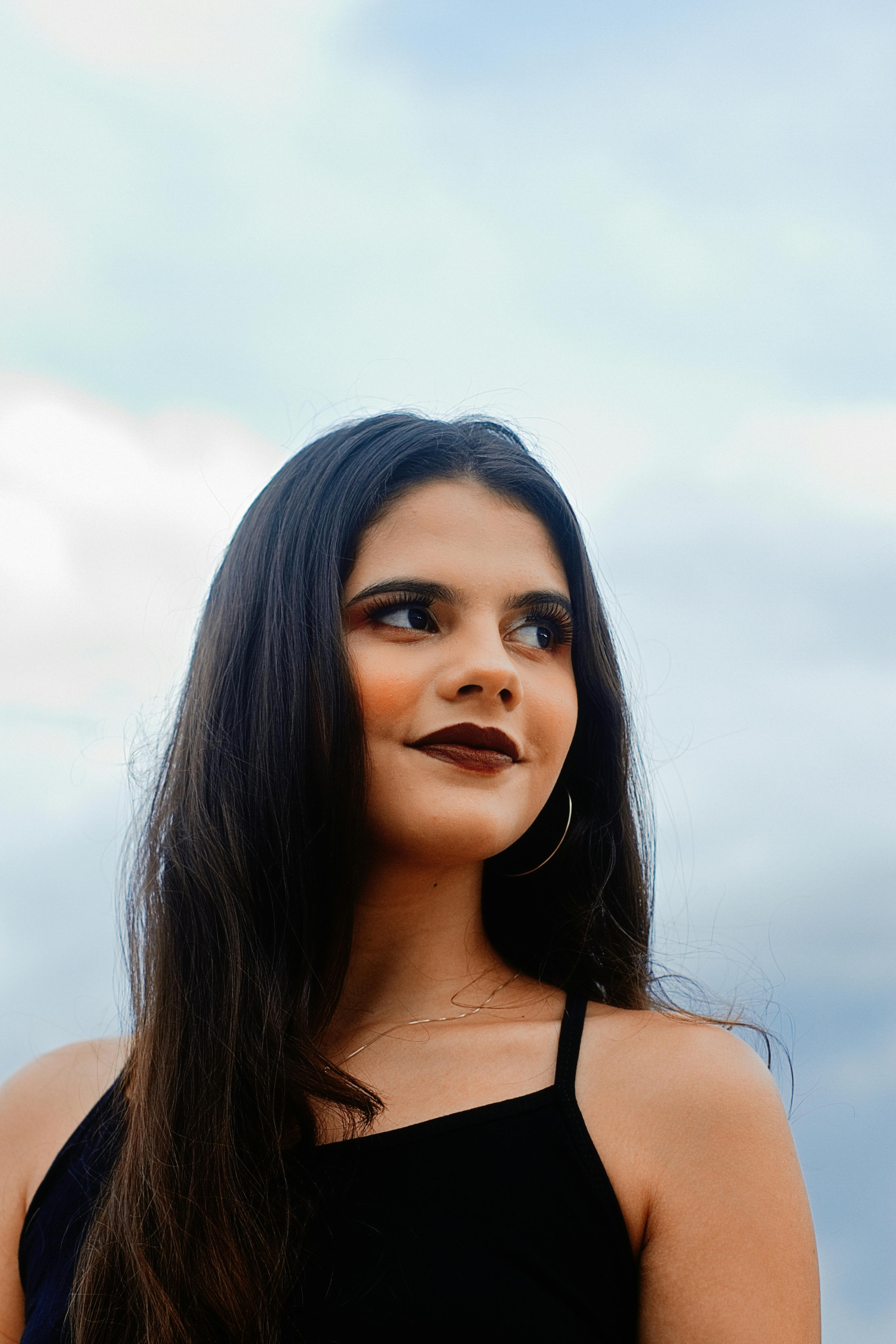 Portrait of a woman with dark lipstick and hoop earrings, gazing away against a cloudy sky backdrop.