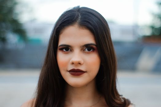 A close-up portrait of a woman with striking makeup, shot outdoors with a blurred background.