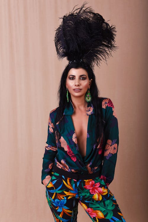 Extravagant female with dark hair in stylish clothes and black feathers on head standing with hands in pockets and looking at camera against beige fabric background