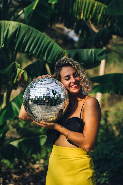 Free A happy woman with blonde curls holds a disco ball amidst lush green foliage, embracing a vibrant outdoor setting. Stock Photo