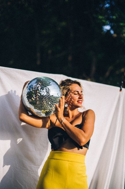 Fashionable woman in vibrant attire holding a disco ball in a sunny outdoor setting.