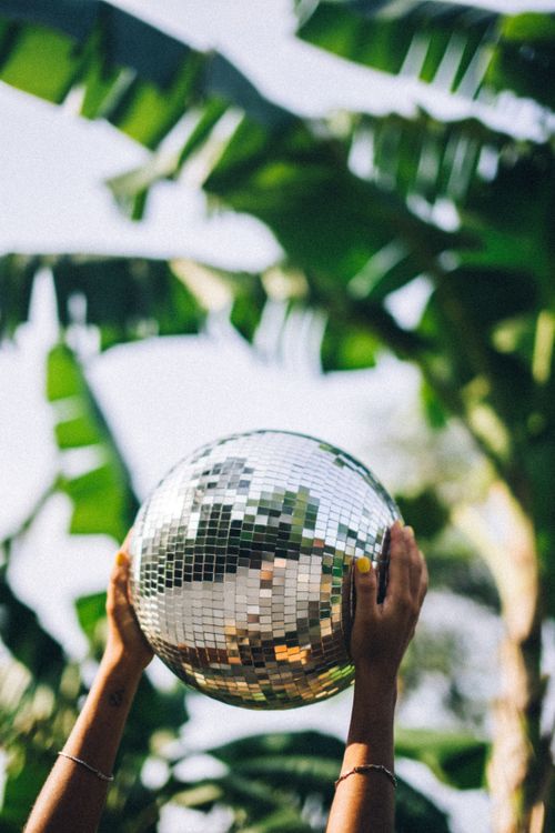 Free A pair of hands hold a disco ball against a backdrop of tropical leaves, capturing a vibrant outdoor scene. Stock Photo
