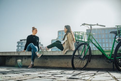 Free Two women enjoy a sunny day sitting outdoors near modern buildings with a bicycle in view. Stock Photo