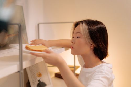 Free A young Asian woman placing a sandwich on a bookshelf, in a cozy indoor setting. Stock Photo