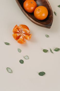 Overhead view of tangerines in wooden bowl with scattered leaves on a beige surface.