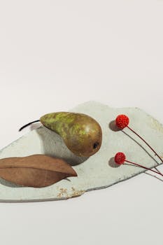 Artistic still life featuring a pear, dried leaf, and red craspedia on a textured surface.