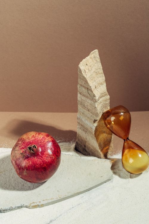 Free Creative still life featuring a pomegranate, sand, stone, and an amber hourglass on a brown background. Stock Photo