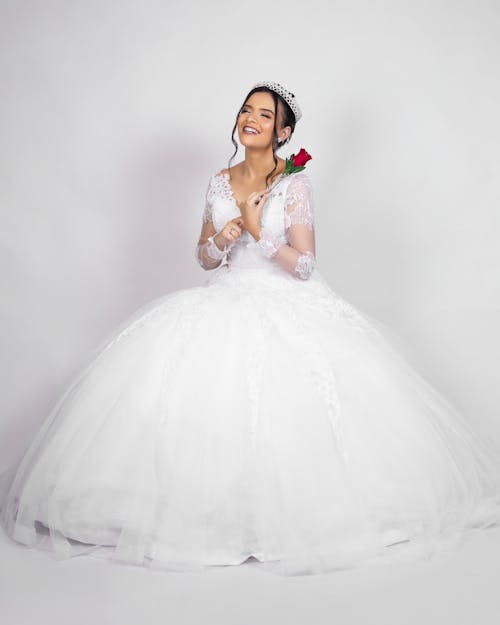 Full body of cheerful young bride in elegant white dress and tiara smiling while standing in light studio with red rose in hand