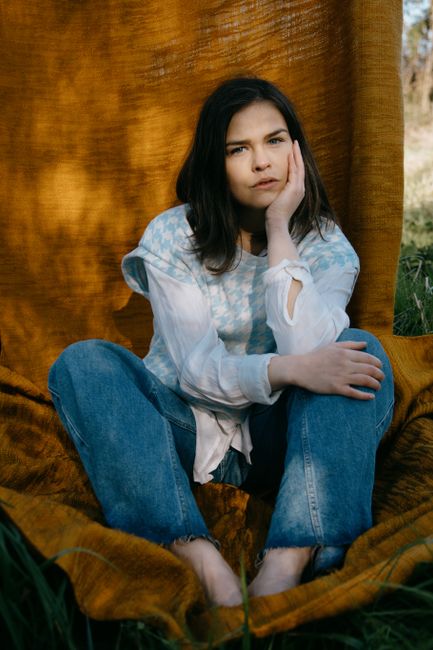 A young woman sitting outdoors in casual clothing against a rustic fabric backdrop.