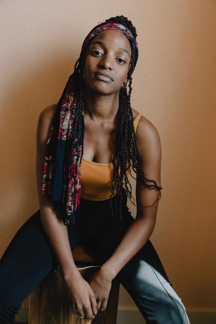 A confident woman in an orange tank top and braided hair poses against a neutral background.