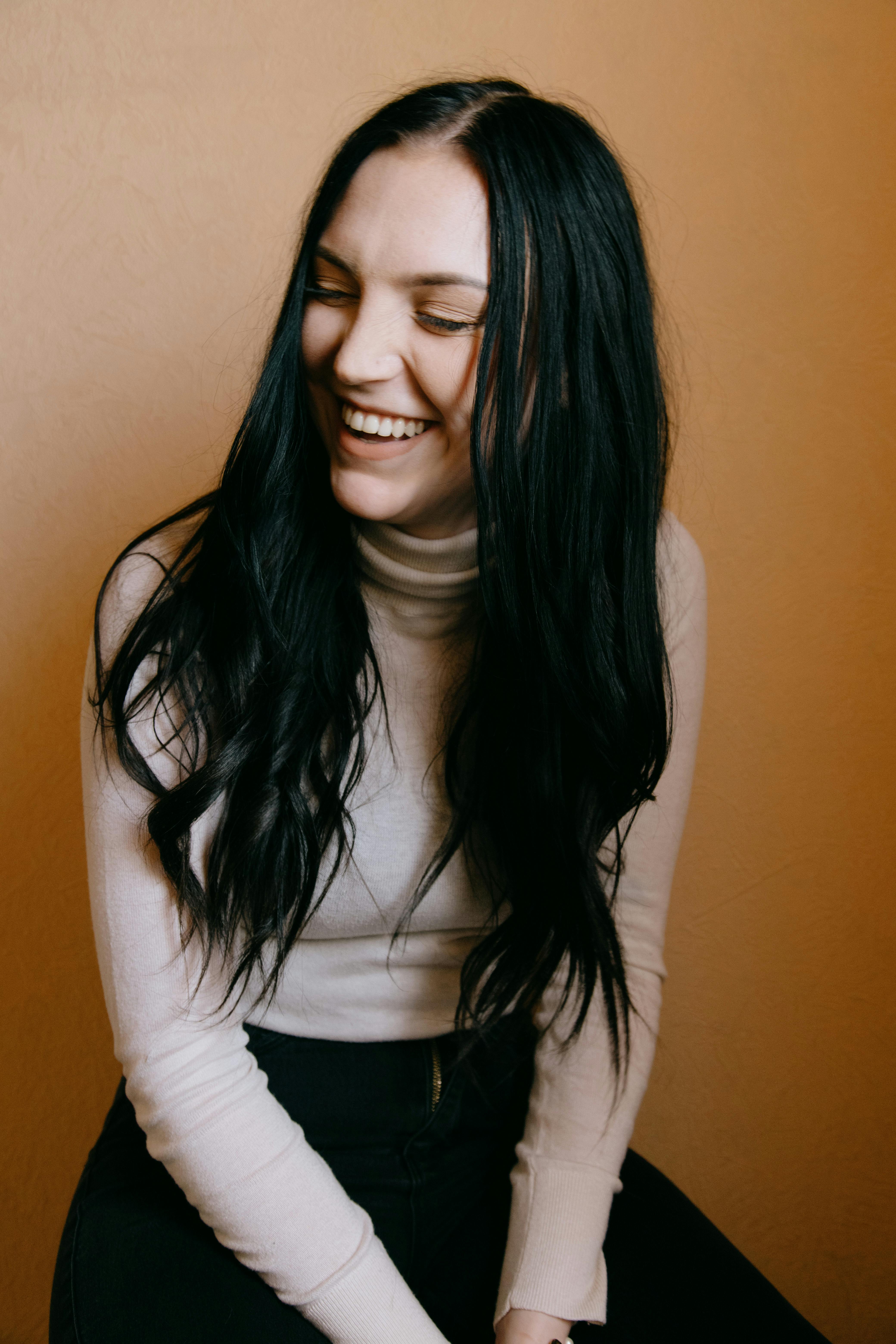 A joyful woman with dark hair laughing while sitting indoors in a white turtleneck.