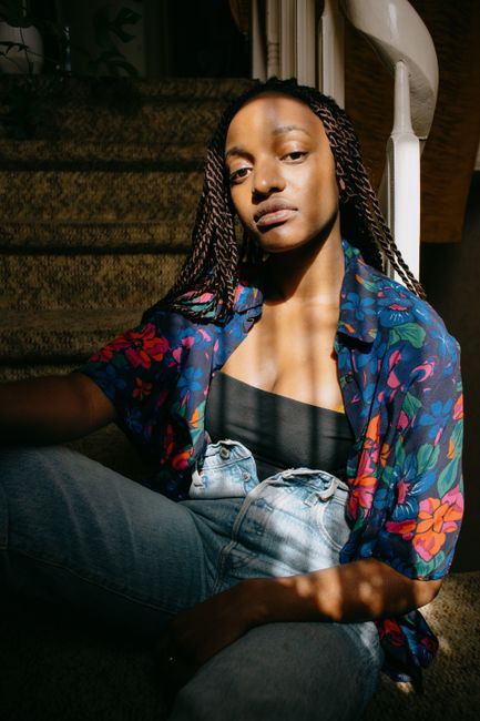 Stylish woman with braids posing on staircase, wearing floral blouse and jeans, illuminated by soft light.