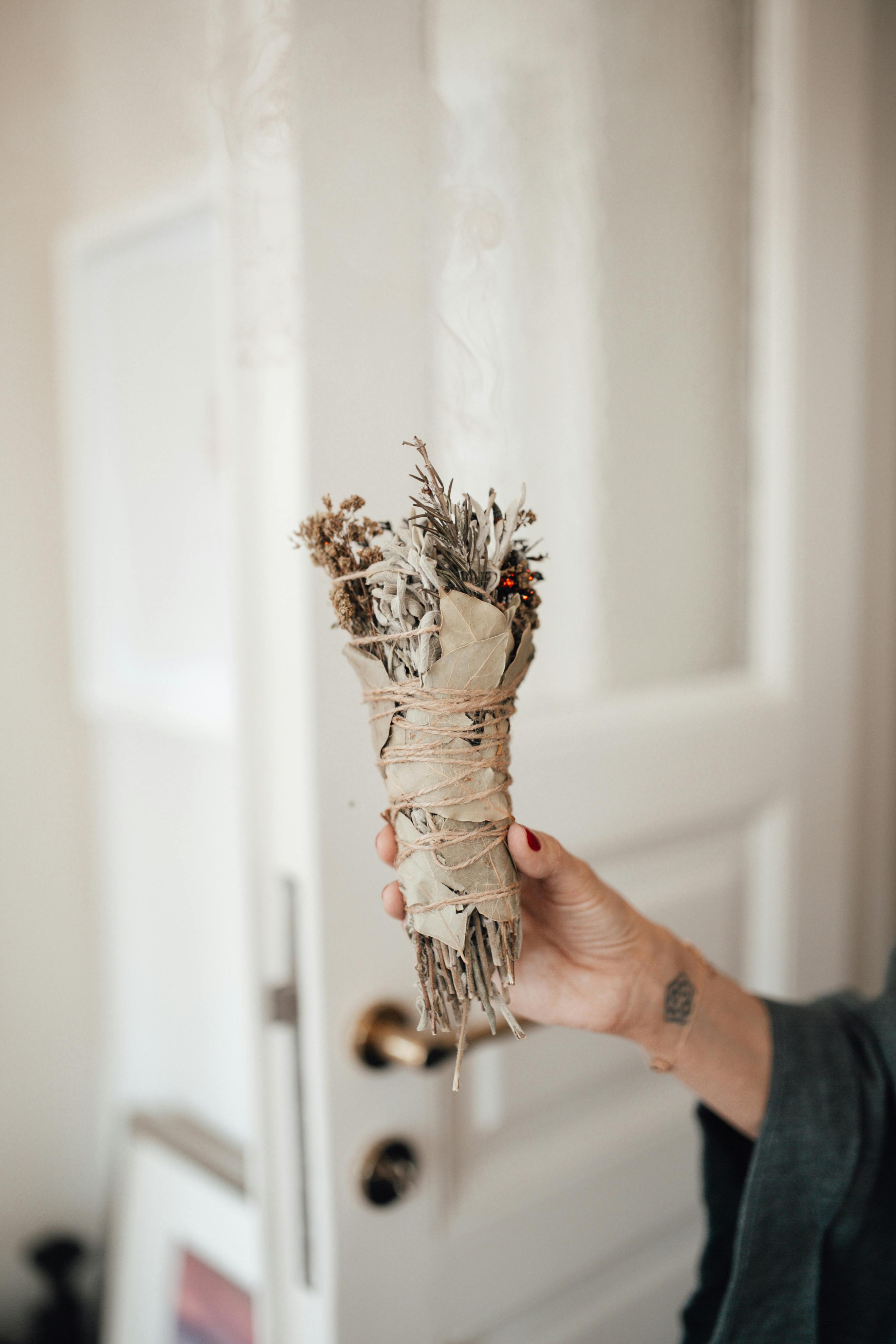 Close-up of a hand holding a sage smudge stick inside a softly lit room.