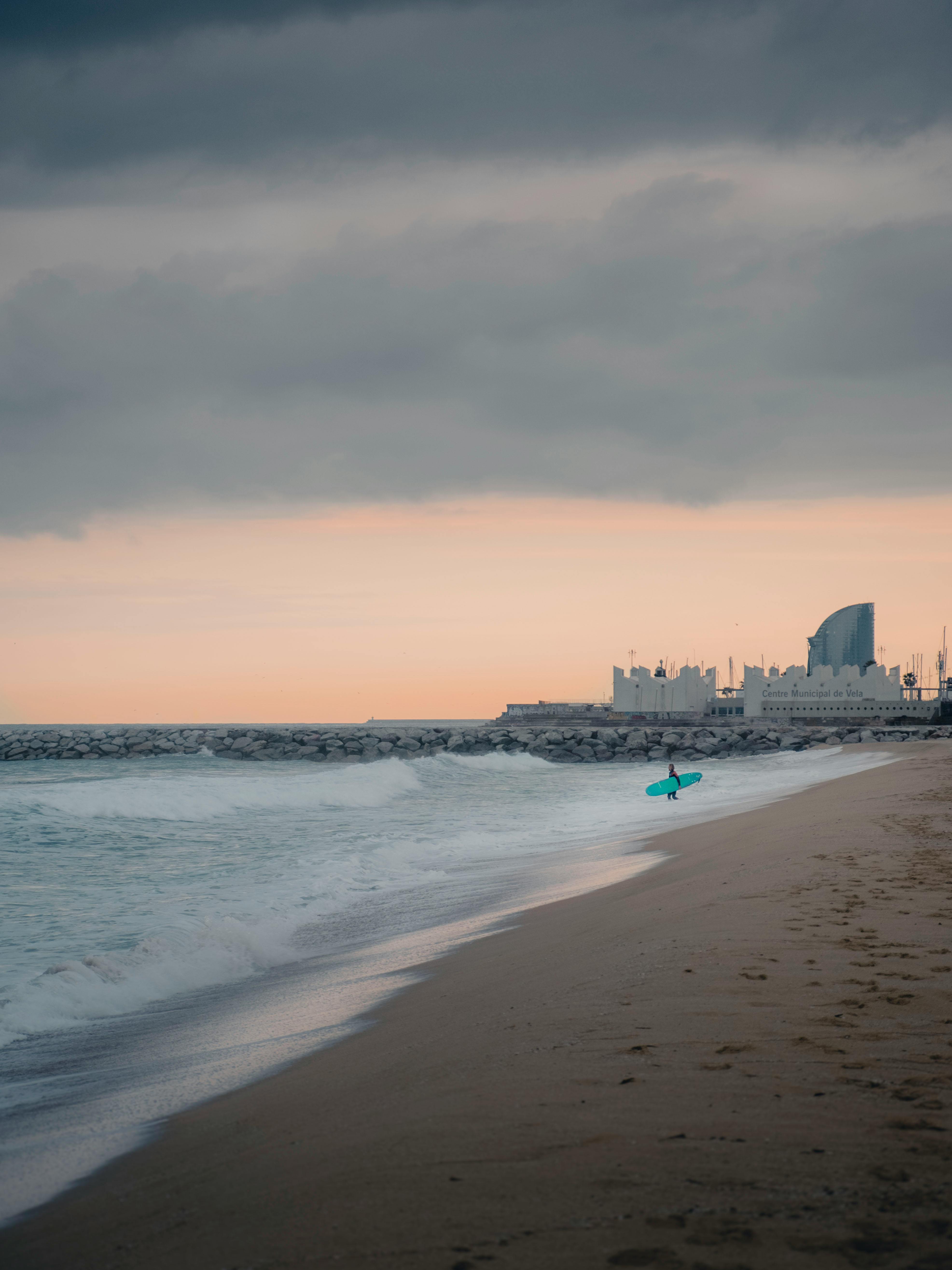 A surfer walks along Barceloneta Beach with waves under a cloudy sunset sky.