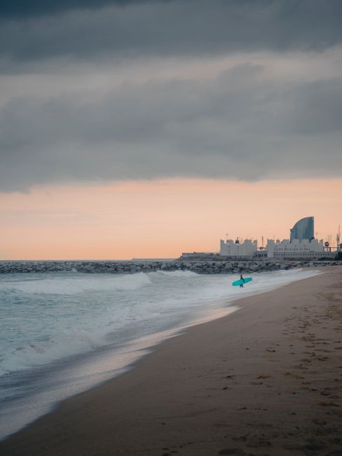 A surfer walks along Barceloneta Beach with waves under a cloudy sunset sky.