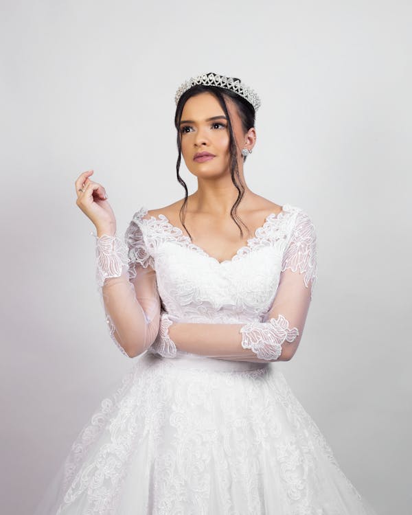 Elegant young bride in classy white dress and tiara standing in studio and looking away dreamily before wedding ceremony