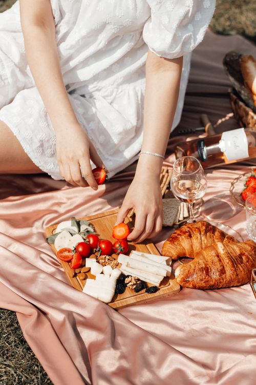 Free A woman's hand setting up a picnic tray with croissants, cheese, and tomatoes on a blanket. Stock Photo