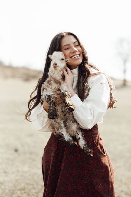 Joyful woman holding a fluffy baby goat, smiling in a sunlit field.